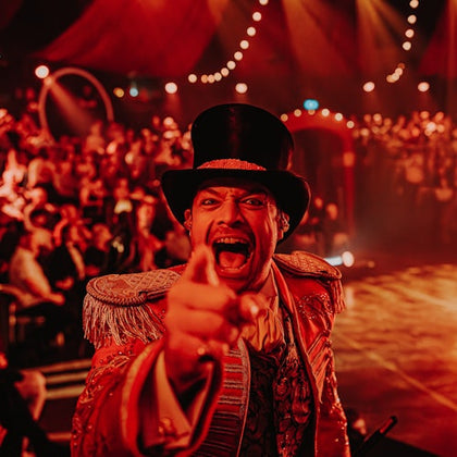 A circus ringmaster in an ornate outfit enthusiastically gestures towards the camera, with a crowd and lights in the background.