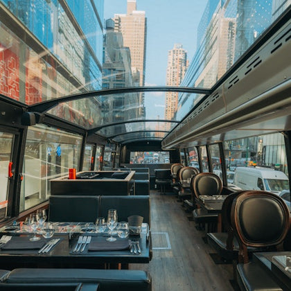 A modern glass-roofed restaurant bus with neatly set tables, parked on a busy city street with tall buildings in the background.
