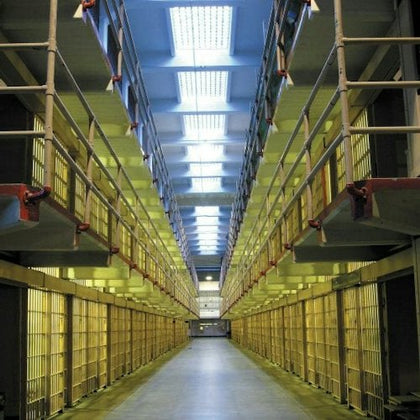 A symmetrical view of a prison hallway flanked by two levels of barred cells, lit by overhead fluorescent lights.