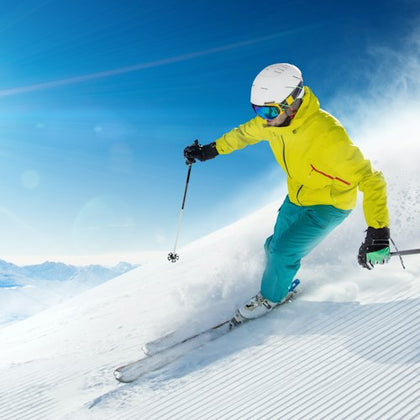 A skier in a yellow jacket and blue pants skiing downhill on a sunny day, leaving a trail of snow behind.