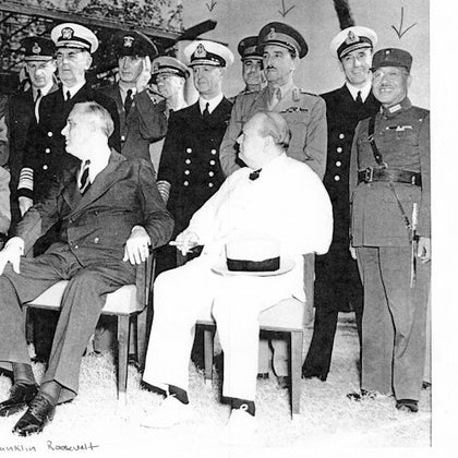 Group of men in military uniforms and suits, with two seated in front and the rest standing behind, outdoors with foliage in the background.