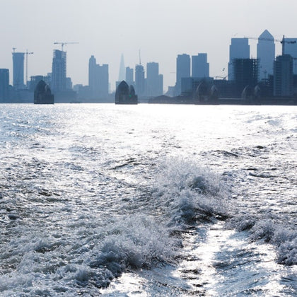 View of a river with waves in the foreground and a cityscape with skyscrapers and cranes in the background.