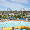 A water park with a wave pool in the foreground, surrounded by shaded seating, with roller coasters in the background.