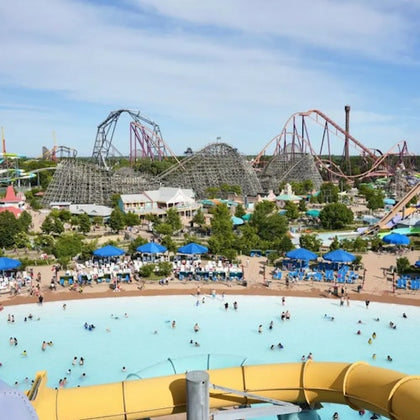 A water park with a wave pool in the foreground, surrounded by shaded seating, with roller coasters in the background.
