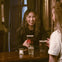 Two women smiling and talking at a pub, one holding a drink on a wooden counter with drinks and a candle.