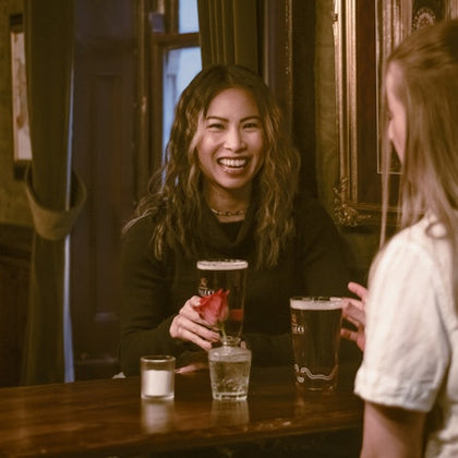 Two women smiling and talking at a pub, one holding a drink on a wooden counter with drinks and a candle.