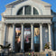 A large neoclassical building with columns, banners, and a large arched window, with people in front on the steps.