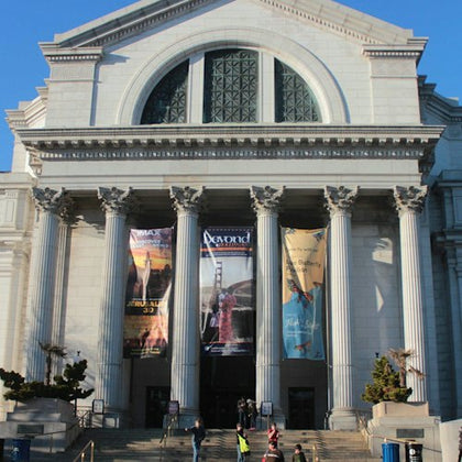 A large neoclassical building with columns, banners, and a large arched window, with people in front on the steps.